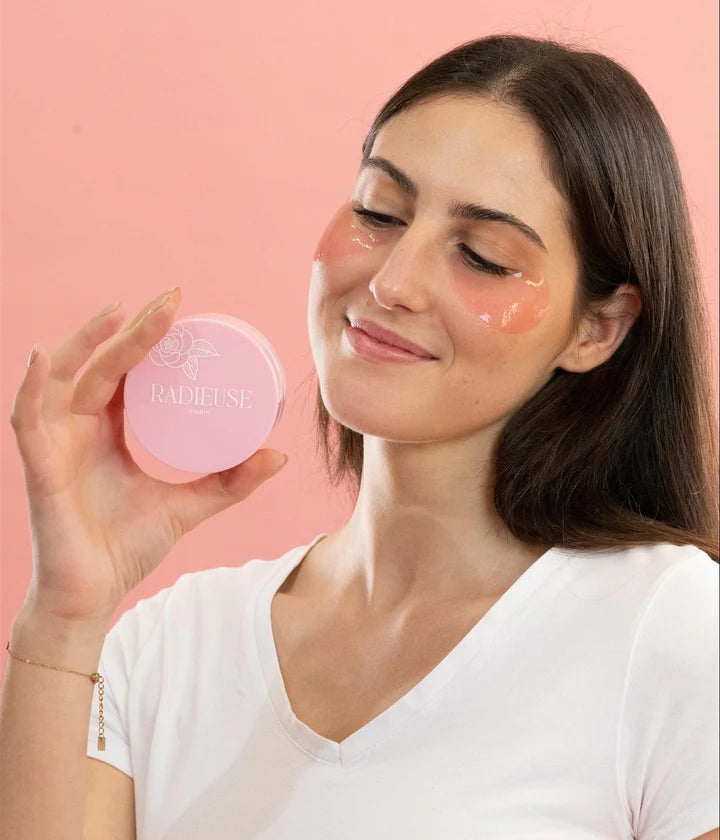 Femme souriante appliquant les patchs anti-cernes Radieuse Paris sous ses yeux, tout en tenant un pot rose du produit. Ces patchs hydratants et décongestionnants au Camélia Japonica aident à réduire les cernes et poches pour un regard lumineux et reposé.