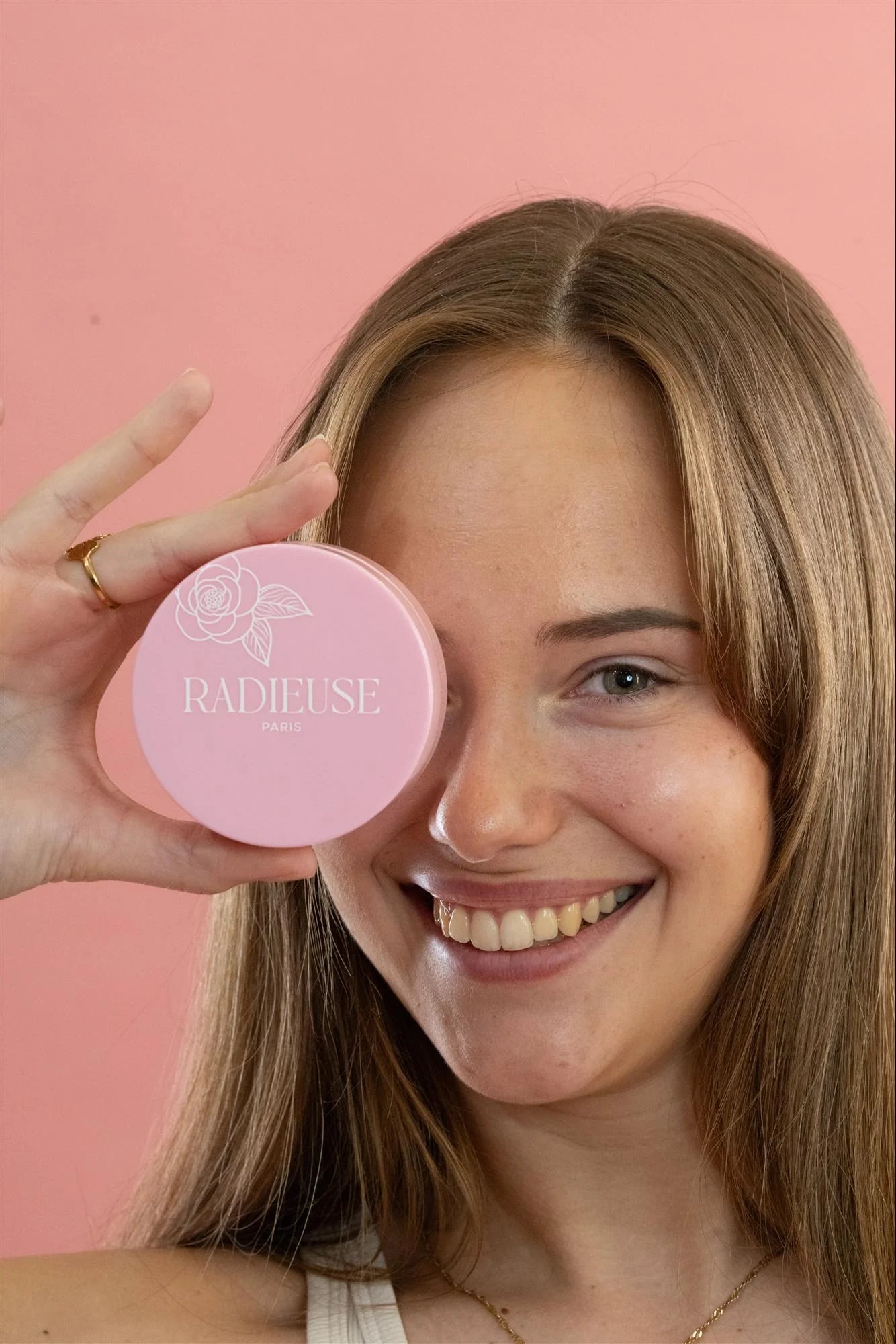 Femme souriante tenant une boîte rose des patchs anti-cernes Radieuse Paris devant son œil. Ce soin au Camélia Japonica hydrate, réduit les cernes et poches, et illumine le regard. Formule vegan et cruelty-free pour un contour des yeux frais et reposé.