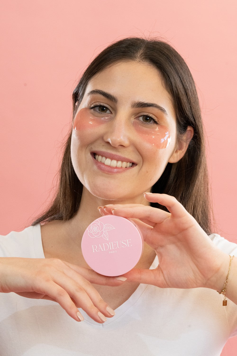 Femme brune souriante portant les patchs anti-cernes Radieuse Paris sous les yeux et tenant une boîte rose du produit. Ces patchs hydrogel hydratants et décongestionnants réduisent les cernes et poches pour un regard éclatant et reposé.