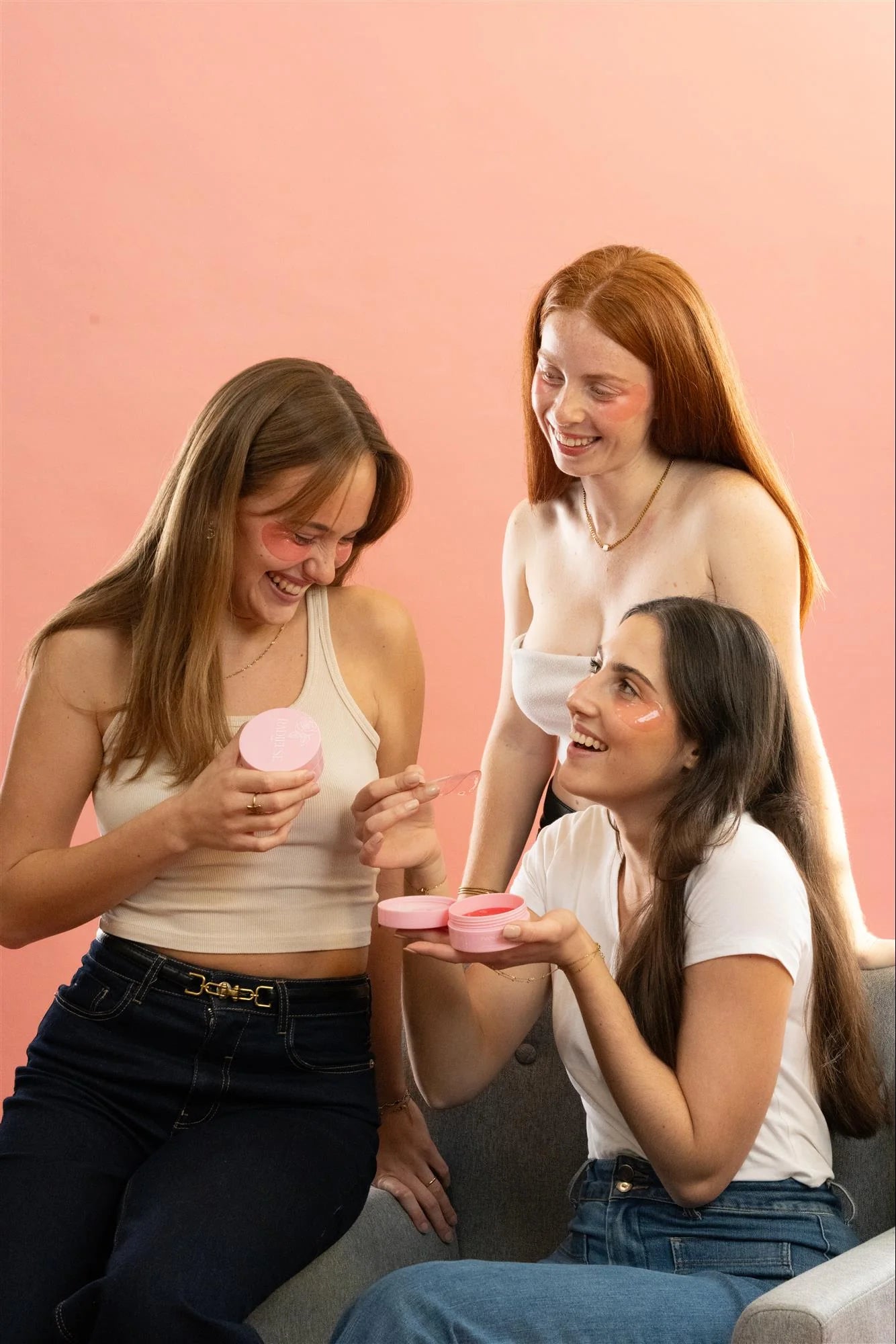 Trois jeunes femmes souriantes appliquant les patchs anti-cernes Radieuse Paris sous leurs yeux. Elles tiennent les boîtes roses du produit et partagent un moment de détente et de complicité. Ces patchs hydratants au Camélia Japonica réduisent les cernes et poches pour un effet fraîcheur instantané.