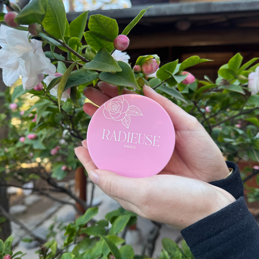 Main tenant un pot rose Radieuse Paris devant une branche de camélia en fleurs – patchs anti-cernes au Camélia Japonica.